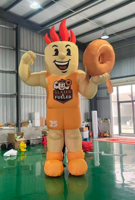 Inflatable mascot in orange basketball uniform holding a donut, flexing its arm, with red hair, in a large, indoor space.