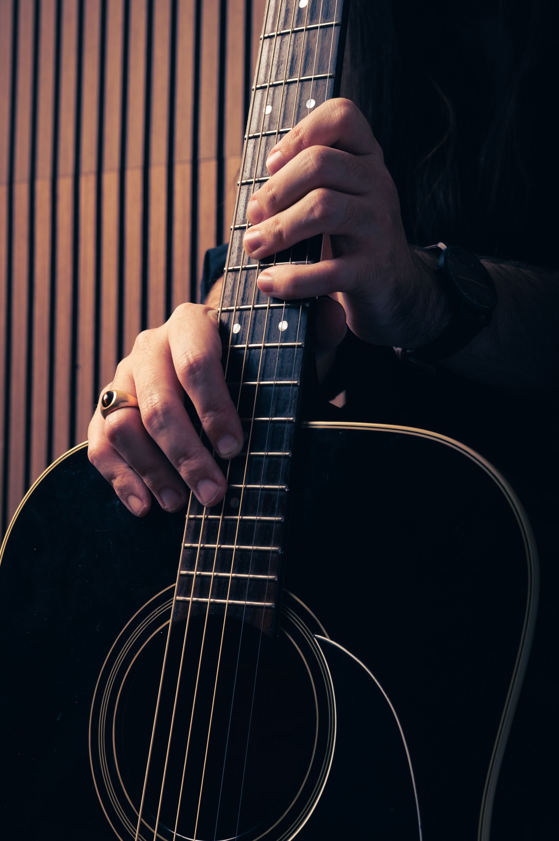 Hands holding the neck of a black acoustic guitar. The setting has a neutral backdrop. Jake Ackerman Music Composer