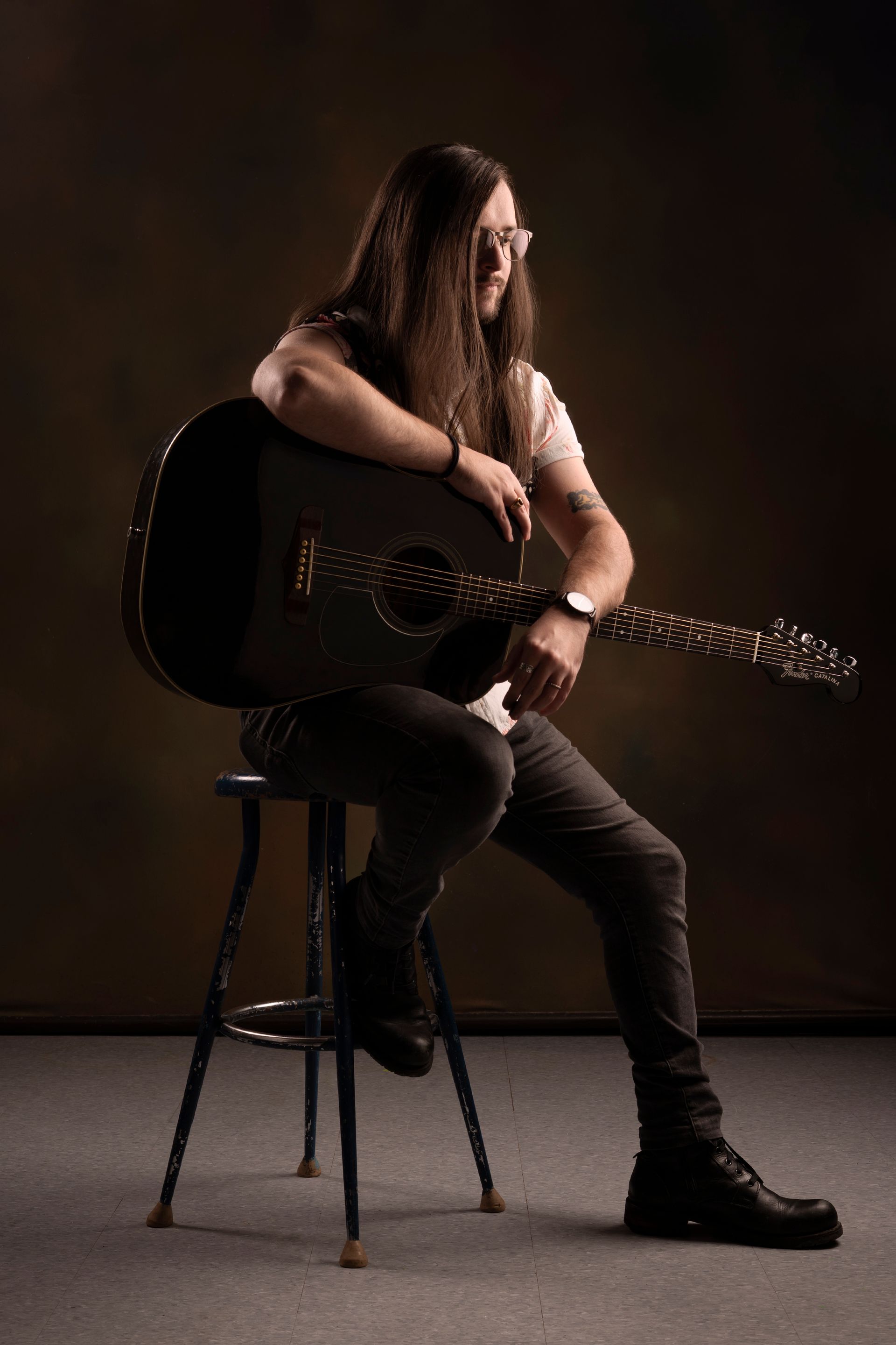 Jake Ackerman with long hair wearing glasses, seated on stool, holding a black acoustic guitar. Dark setting. Jake Ackerman Music, Composer for video games, movies, and digital media