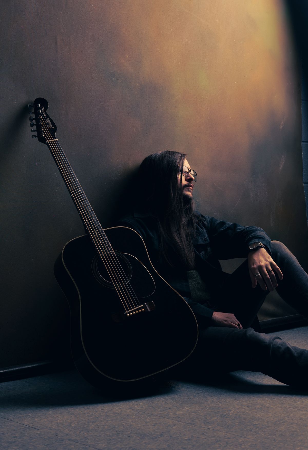 Man with long hair sits leaning against a wall, next to a black acoustic guitar. Image of Jake from Jake Ackerman music