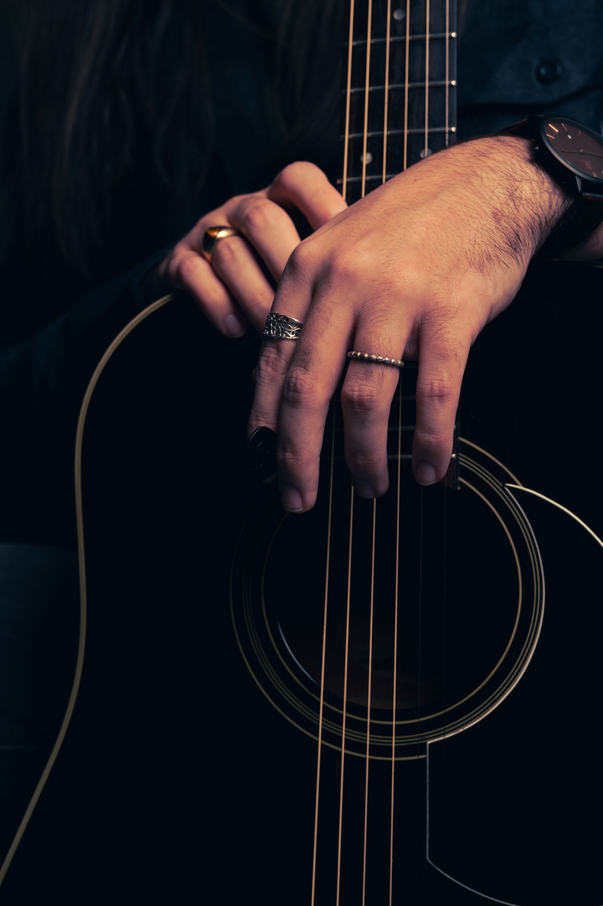 Hands with rings resting on the strings of a black acoustic guitar.