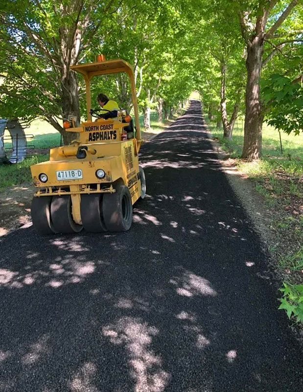 A Yellow Road Roller is Driving Down a Road Surrounded by Trees — North Coast Asphalts in Corindi Beach, NSW