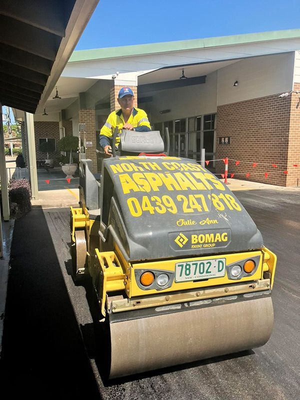 A Man is Driving a Yellow and Black Asphalt Roller in Front of a Building — North Coast Asphalts in Corindi Beach, NSW