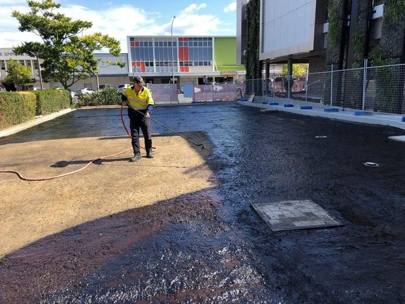 A Man is Spraying Asphalt in a Parking Lot With a Hose — North Coast Asphalts in Corindi Beach, NSW