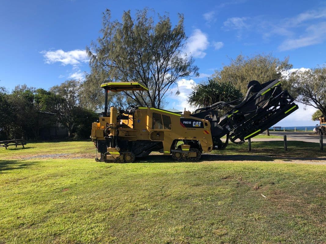 Yellow Construction Vehicle on Grass, Black Contraption Attached, Trees and Ocean in Background — North Coast Asphalts in Corindi Beach, NSW