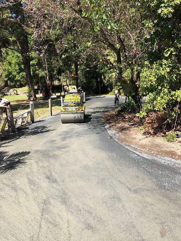 A Yellow Road Roller is Driving Down a Road Surrounded by Trees — North Coast Asphalts in Corindi Beach, NSW