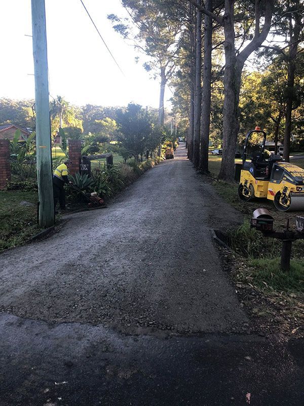 A Yellow Tractor is Driving Down a Road Next to Trees — North Coast Asphalts in Corindi Beach, NSW