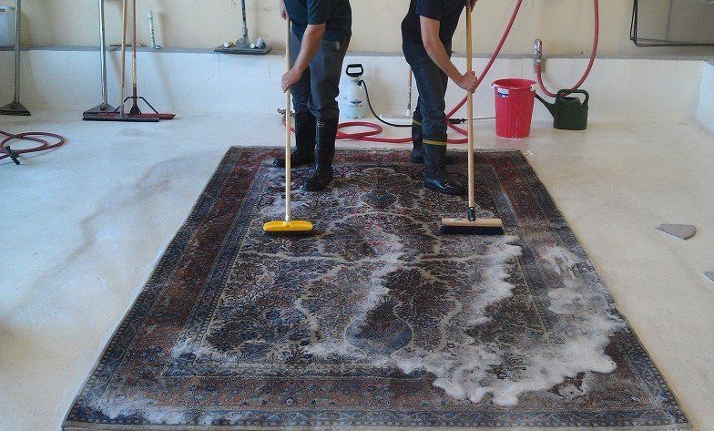 Two men are cleaning a rug with brooms