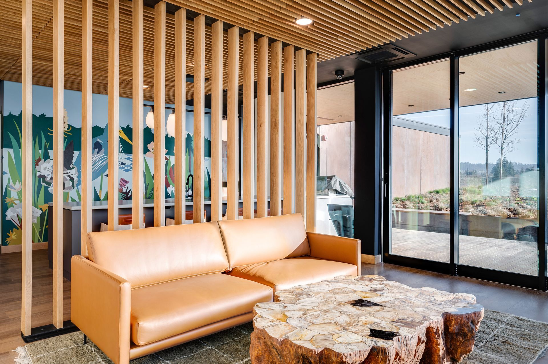 Living room with tan leather sofa, wooden slat wall, and stone coffee table. Large windows overlook a landscape.