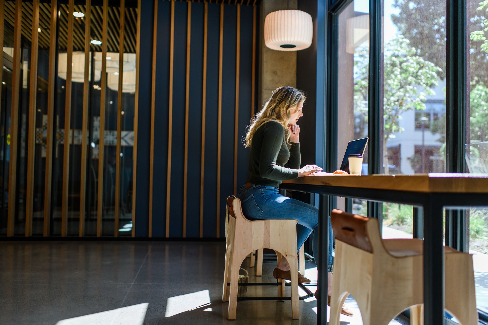 Woman works on laptop at a table by a large window; interior office space.