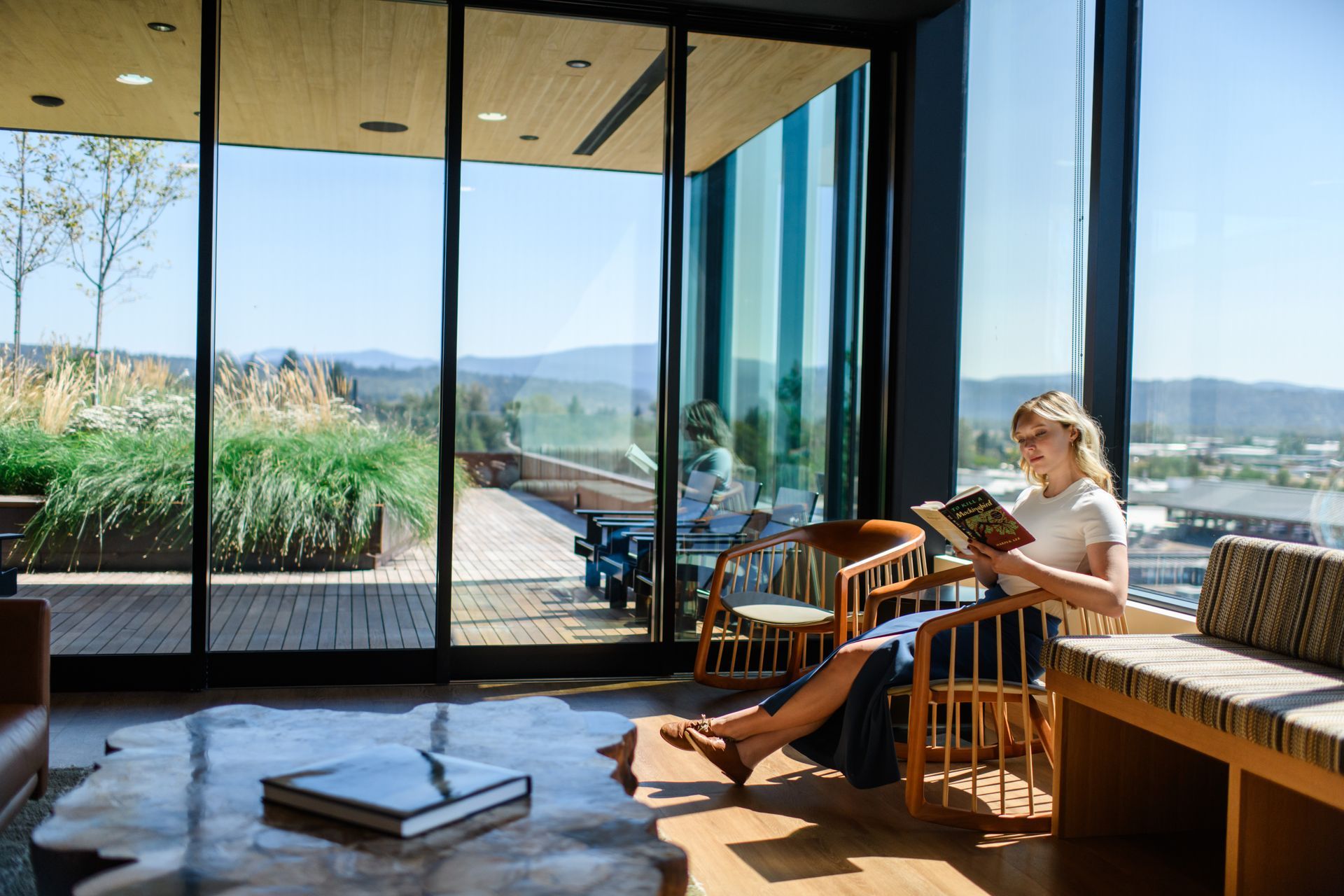 Woman reads a book in a modern room with a large window overlooking a landscape on a sunny day.