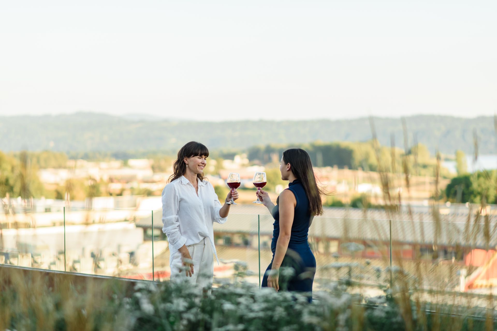 Two people toasting with wine glasses on a rooftop overlooking a city and hills.