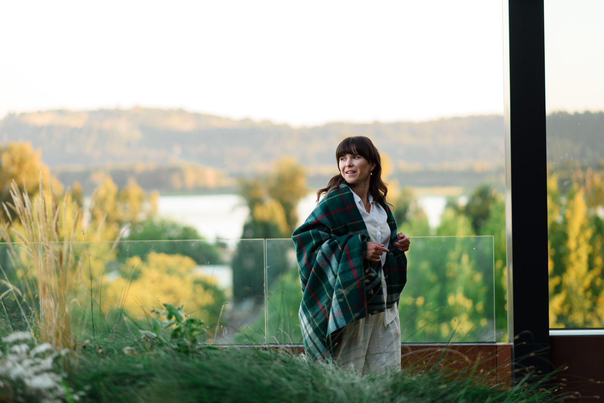 Woman wrapped in green shawl, standing on a balcony overlooking a lake and trees.