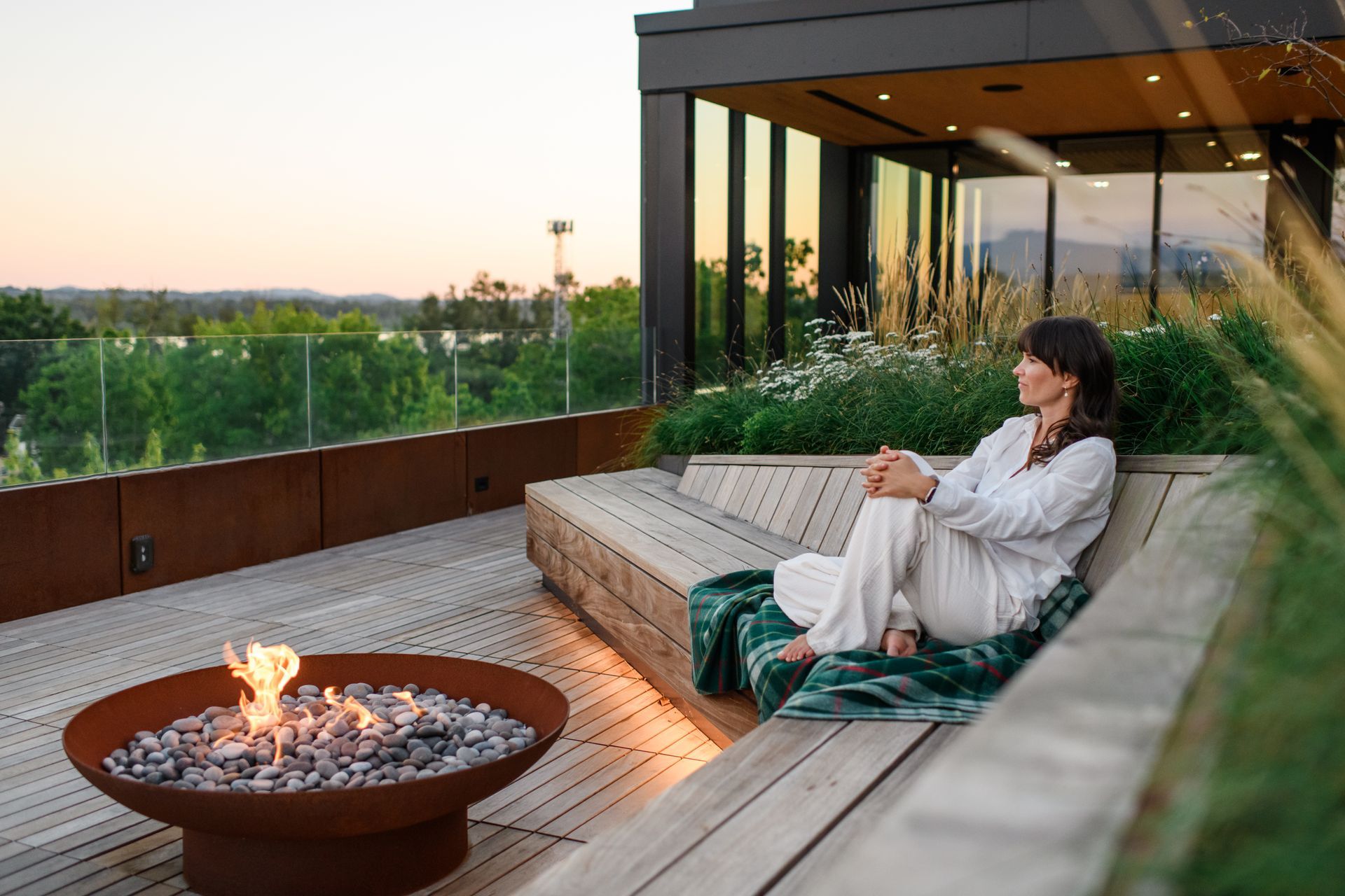 Woman sitting by a fire pit on a rooftop deck, looking out at the view.