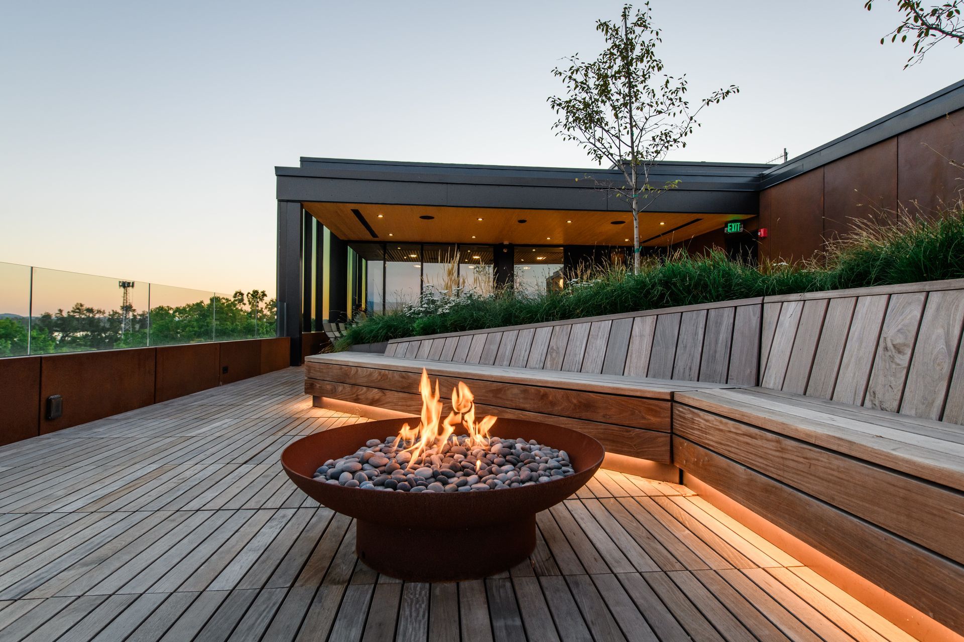 Outdoor rooftop with fire pit, wooden benches, and city view at dusk.