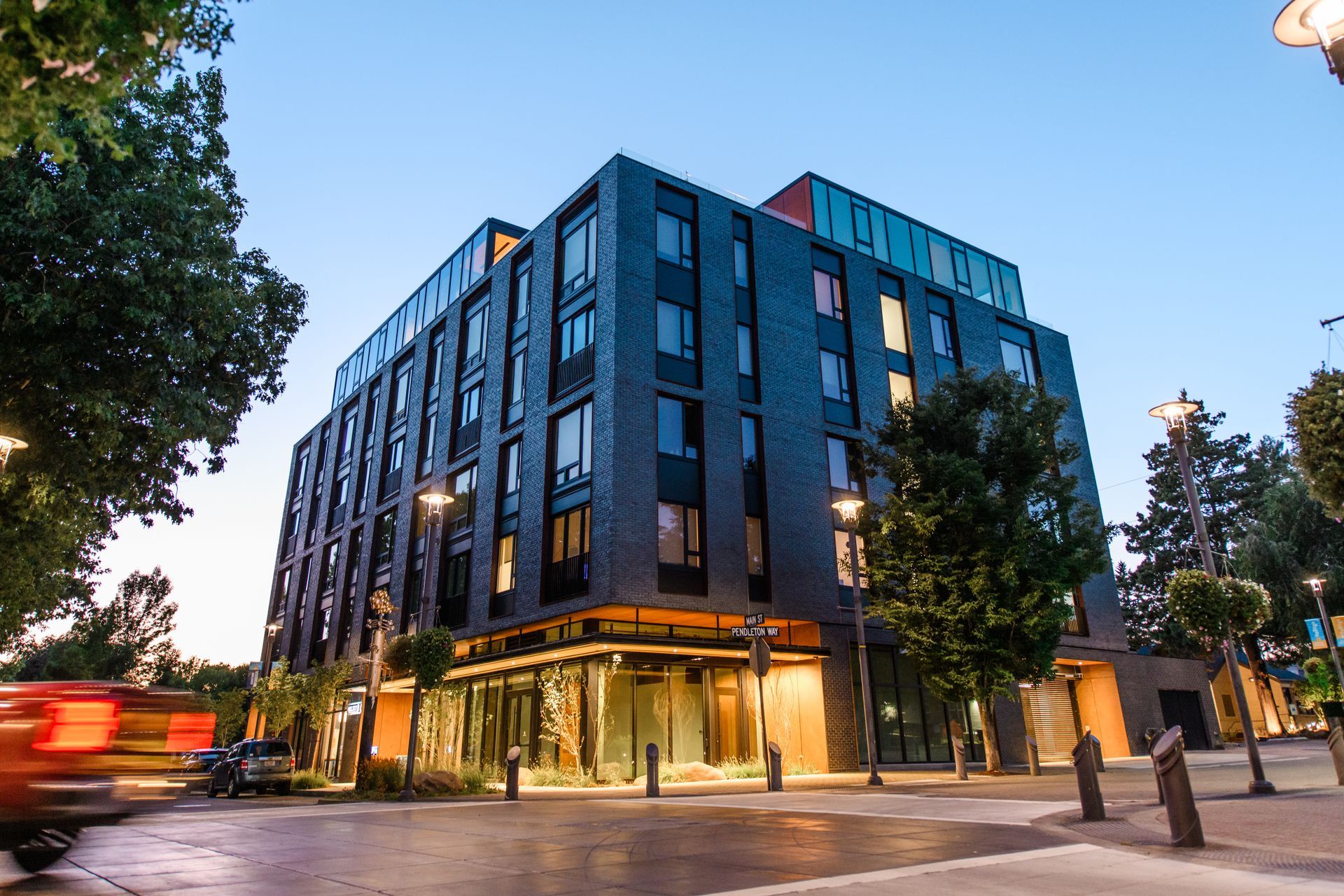 Modern multi-story building at dusk with illuminated ground-level storefronts; street lights and passing car.