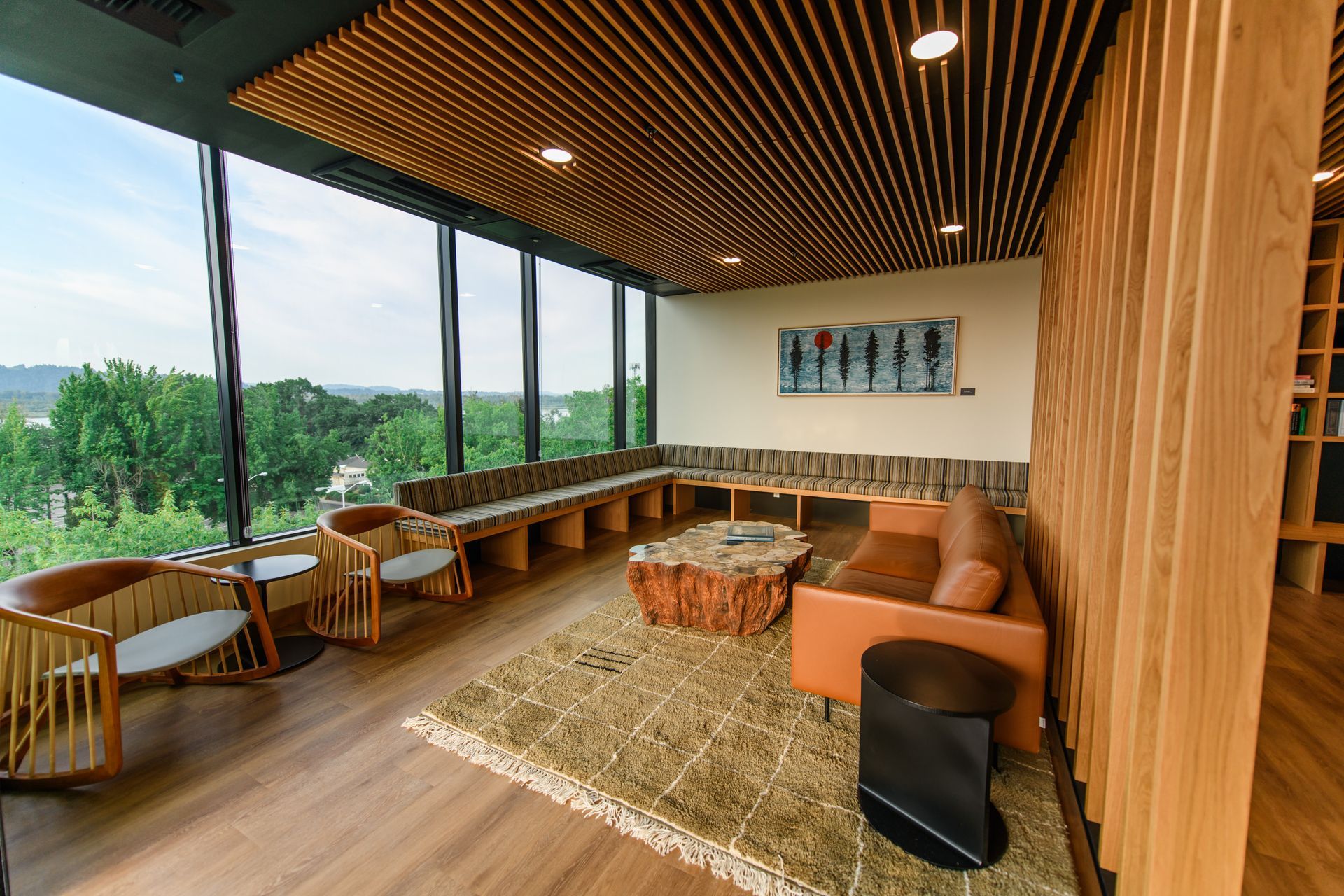 Modern lounge with floor-to-ceiling windows, wood accents, and a view of greenery. Furnishings include chairs and a sofa.