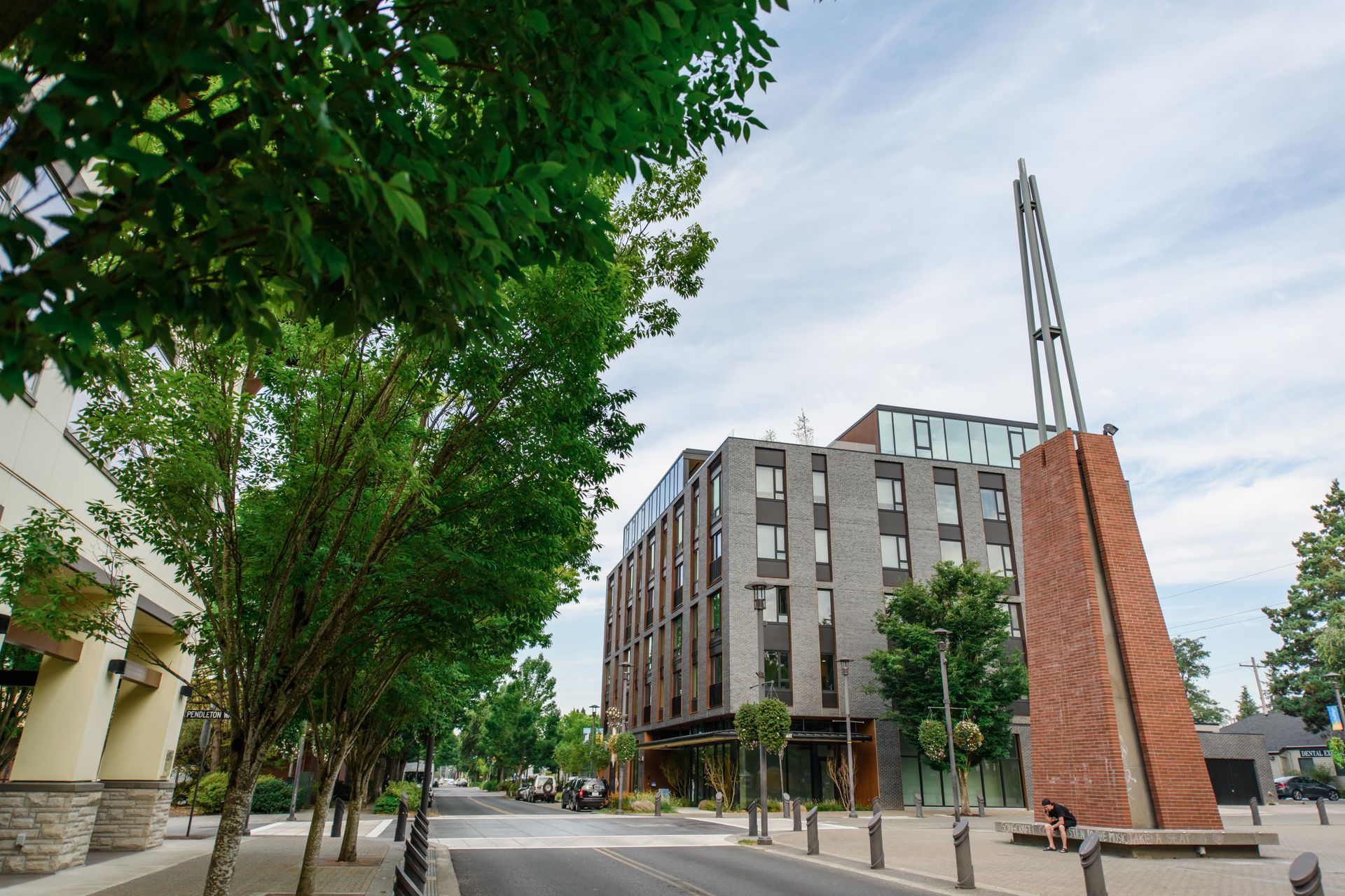 Street view with a modern brick and glass building, a reddish-brown sculpture, and trees under a cloudy sky.