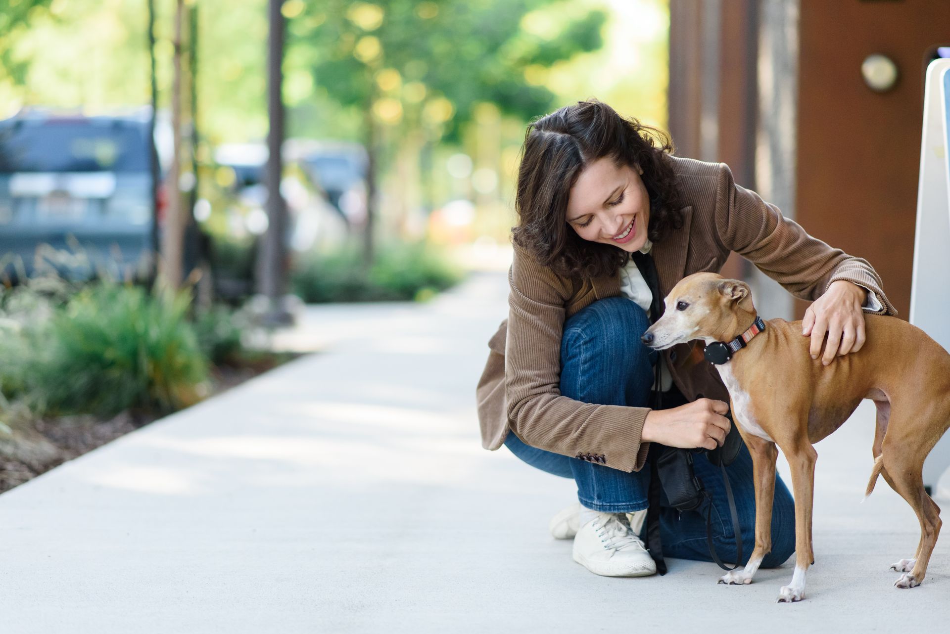 Woman kneeling on a sidewalk, petting a small brown dog; trees and building in the background.
