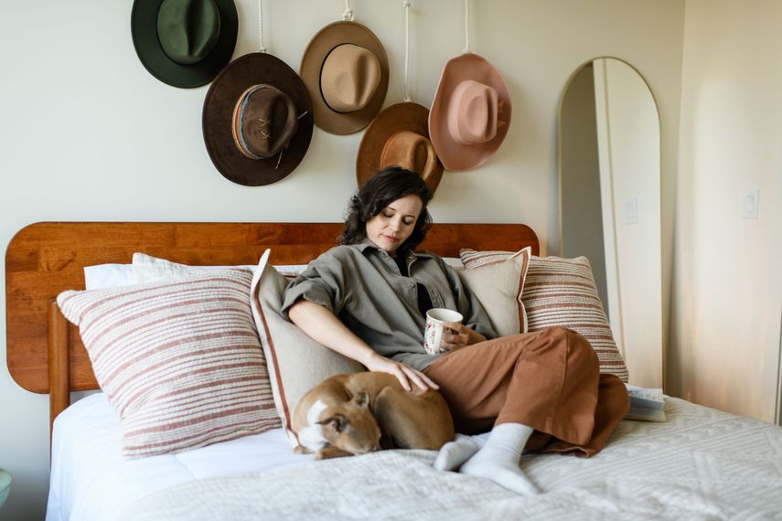 Woman petting dog on bed, holding mug, surrounded by pillows. Hats hang above.