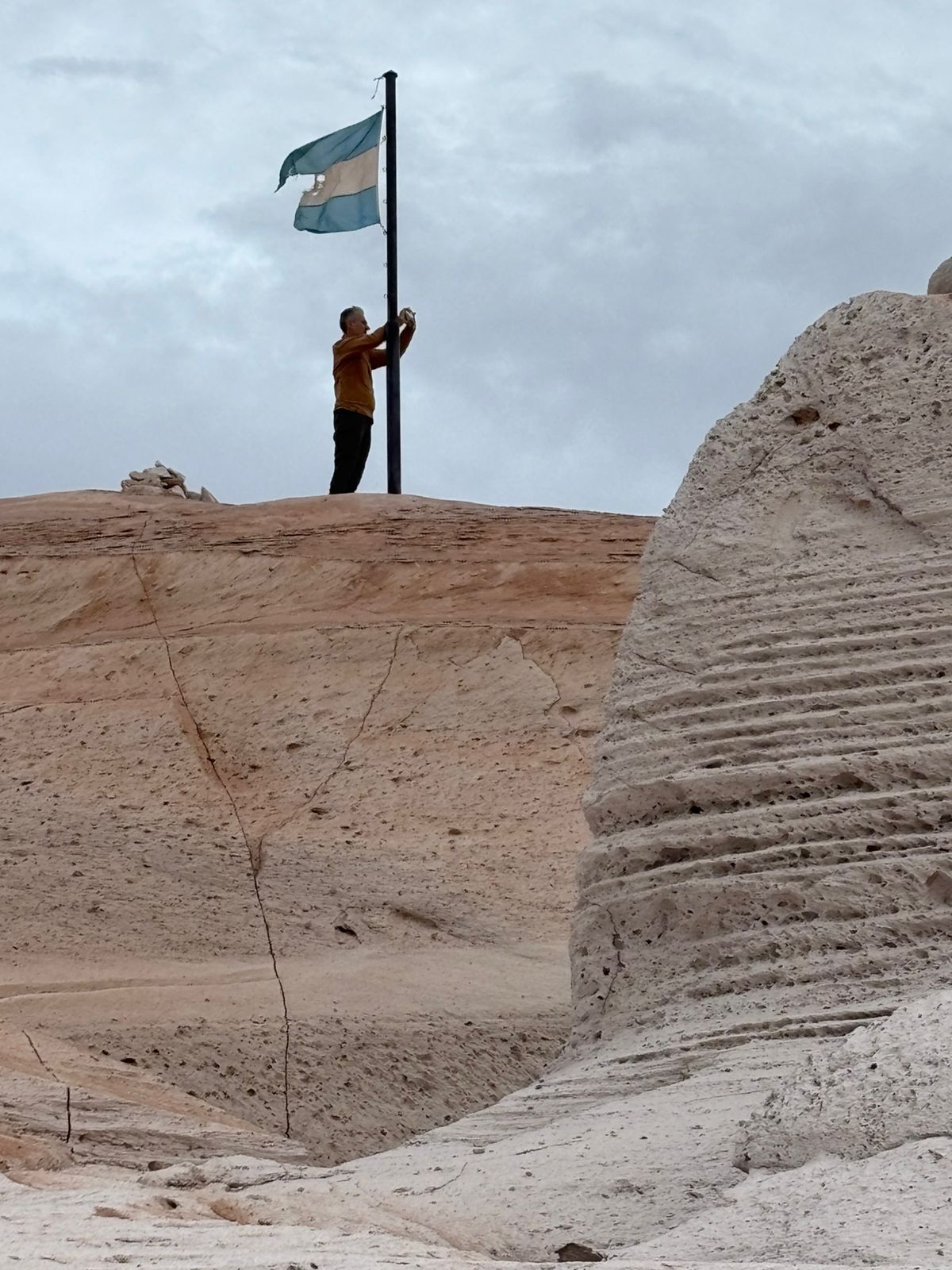 Un hombre iza la bandera argentina en la cima de una colina con formaciones rocosas blancas en capas bajo un cielo nublado.