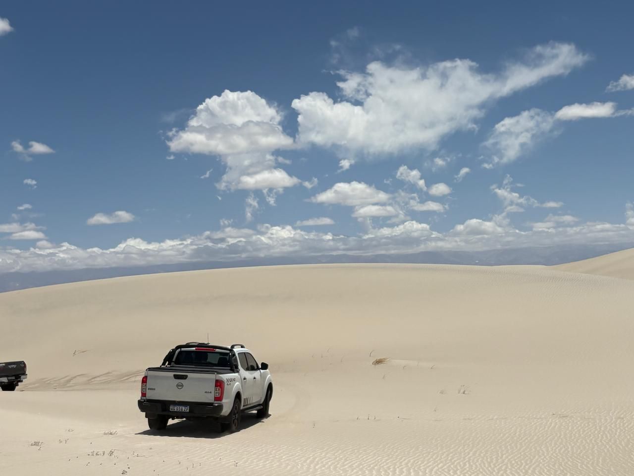 Una camioneta blanca circula por un vasto desierto de arena bajo un cielo azul con esponjosas nubes blancas.