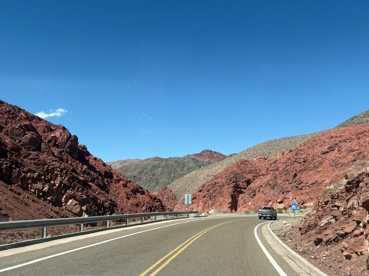 La carretera serpentea a través de montañas de roca roja bajo un cielo azul brillante, con un automóvil conduciendo.