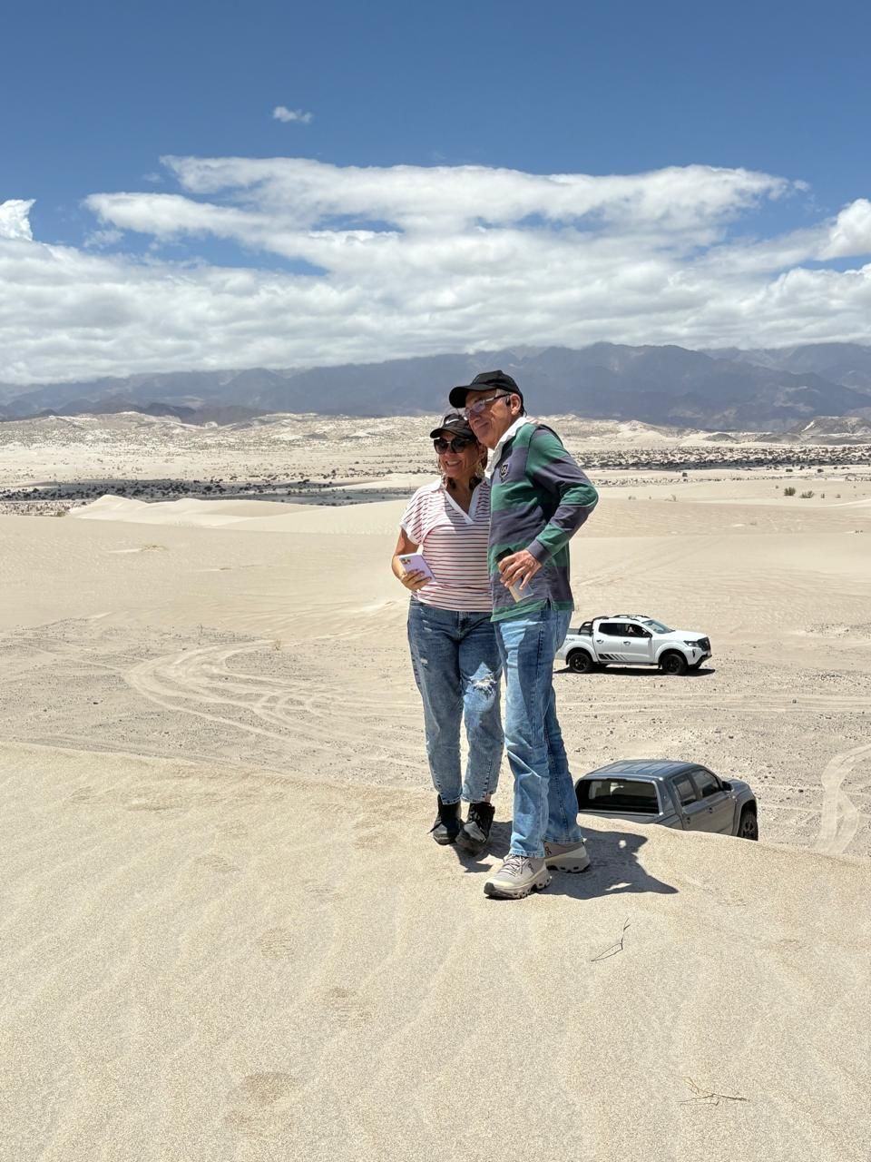 Pareja de pie sobre una duna de arena con un automóvil visible al fondo contra un fondo de montañas y un cielo azul.