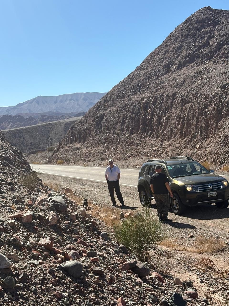 Dos hombres en una camioneta marrón por una carretera desértica con una gran formación rocosa al fondo. Día soleado.