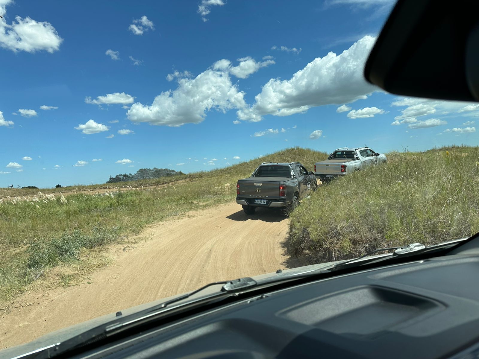 Dos camiones circulando por un camino de tierra a través de un campo de hierba bajo un cielo azul con nubes.