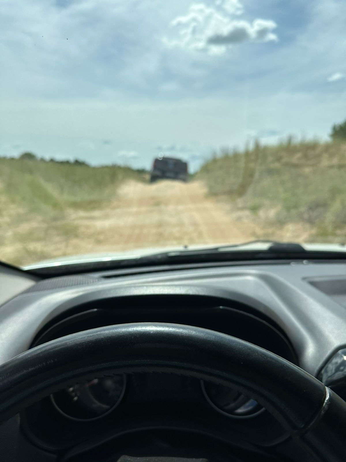 Vista desde el interior de un vehículo circulando por un camino de tierra hacia otro vehículo a lo lejos. Cielo nublado.