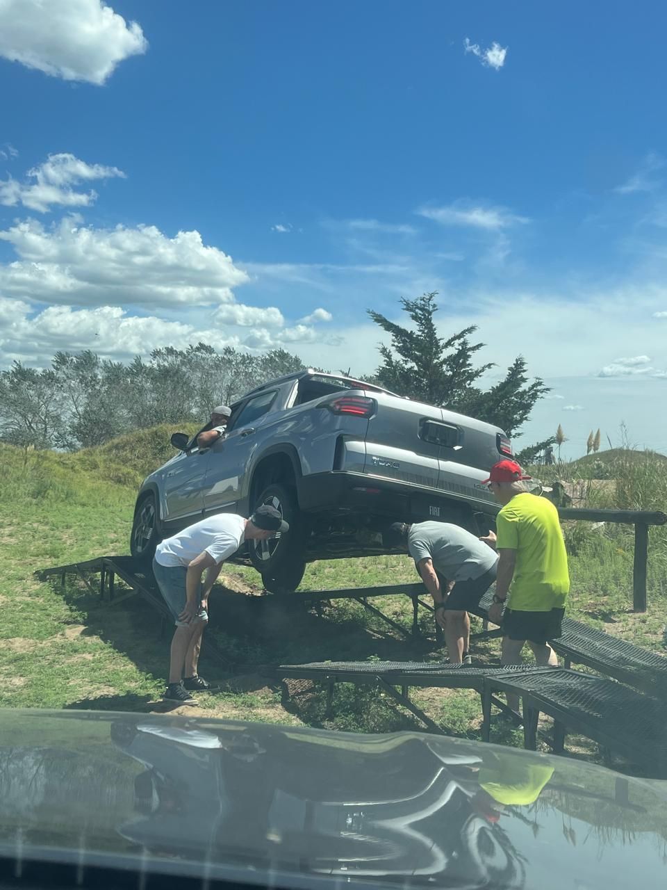 Tres personas inspeccionando la parte inferior de un automóvil en una rampa, en un entorno exterior soleado.