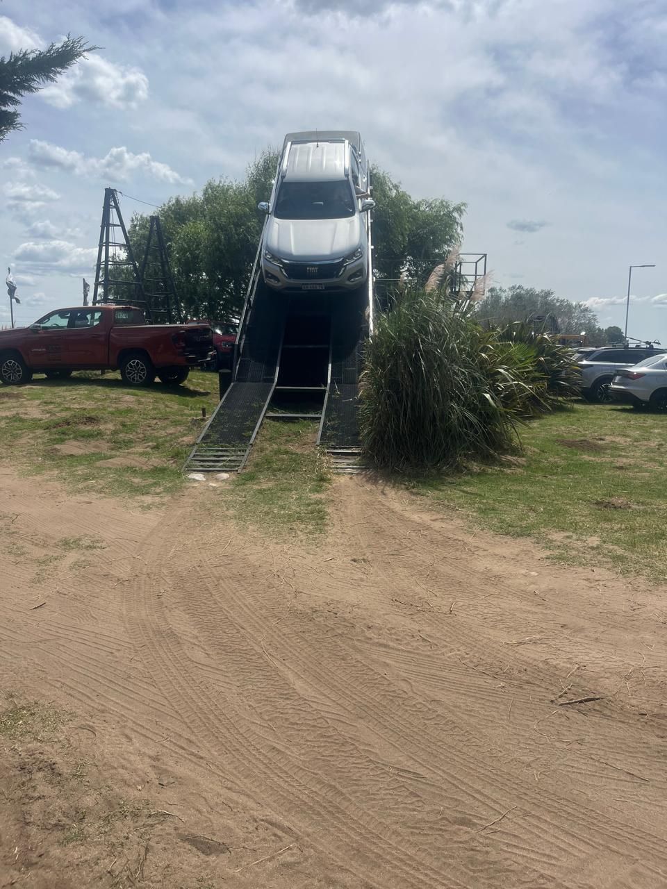 SUV en una rampa, estacionada sobre una plataforma elevada. La rampa baja a un terreno arenoso; camioneta roja al fondo.