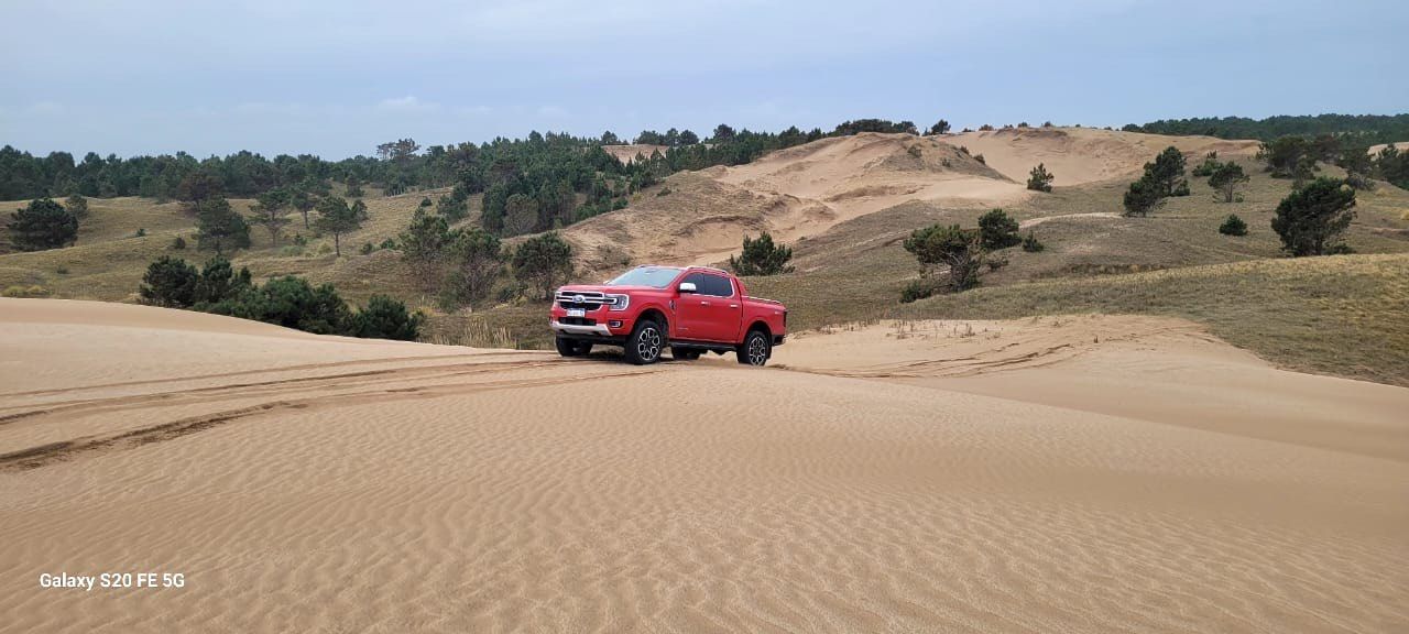 Una camioneta roja circula por una duna de arena. Al fondo, árboles y colinas bajo un cielo nublado.