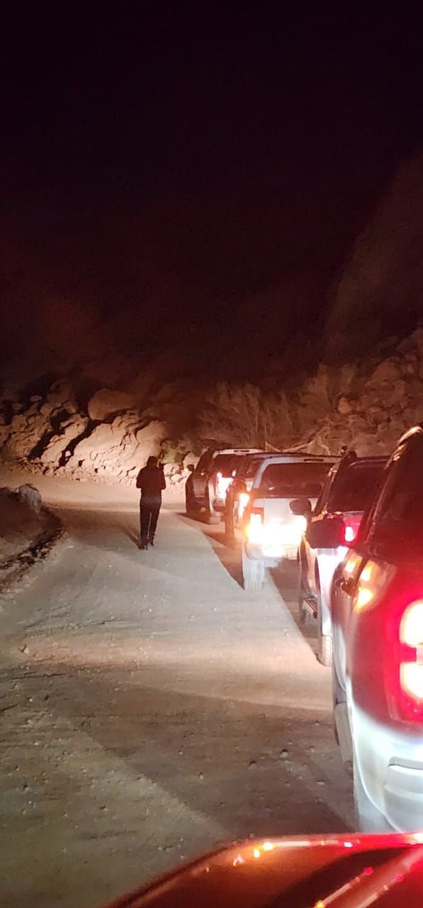 Una persona camina de noche por un camino estrecho y oscuro, bordeado de coches aparcados y una ladera rocosa.