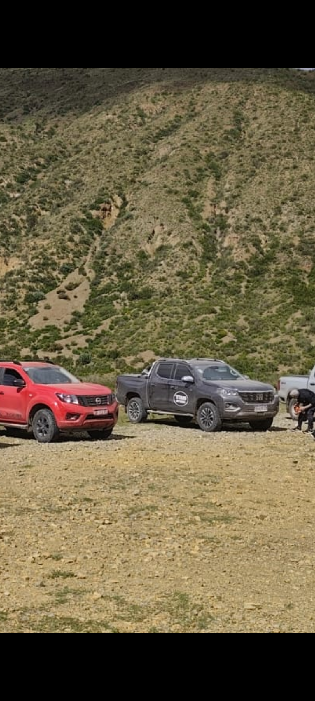 Un camión rojo y una camioneta gris estaban estacionados en una pendiente seca y rocosa al pie de una ladera verde.