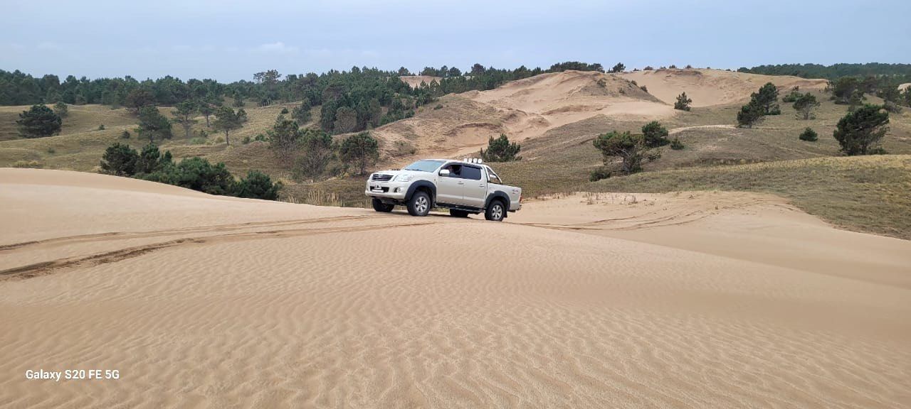 Una camioneta blanca circula por una duna de arena, con un fondo de otras dunas y árboles verdes bajo un cielo nublado.