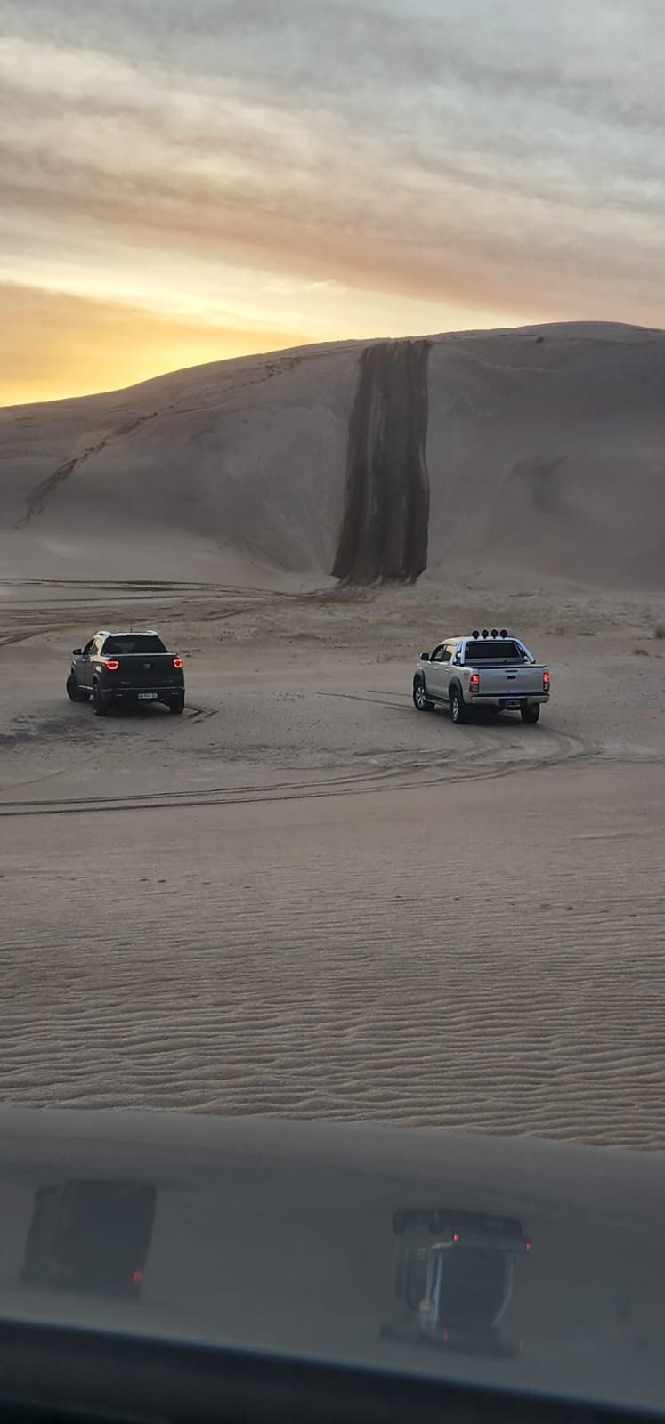Dos vehículos sobre la arena con una gran duna al fondo bajo un cielo de atardecer.