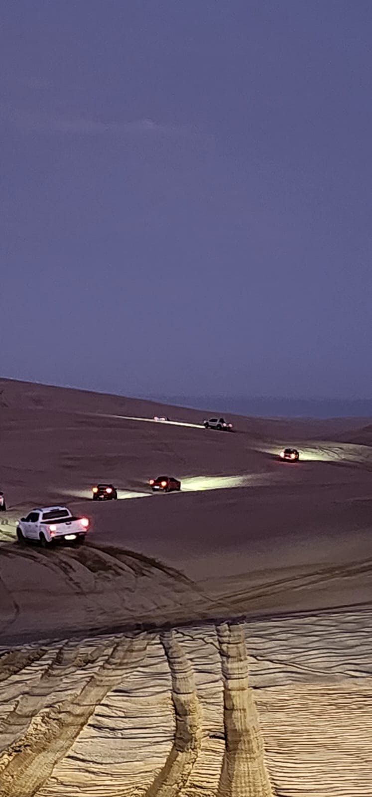Coches circulando por un paisaje desértico al anochecer, luces iluminando la arena.