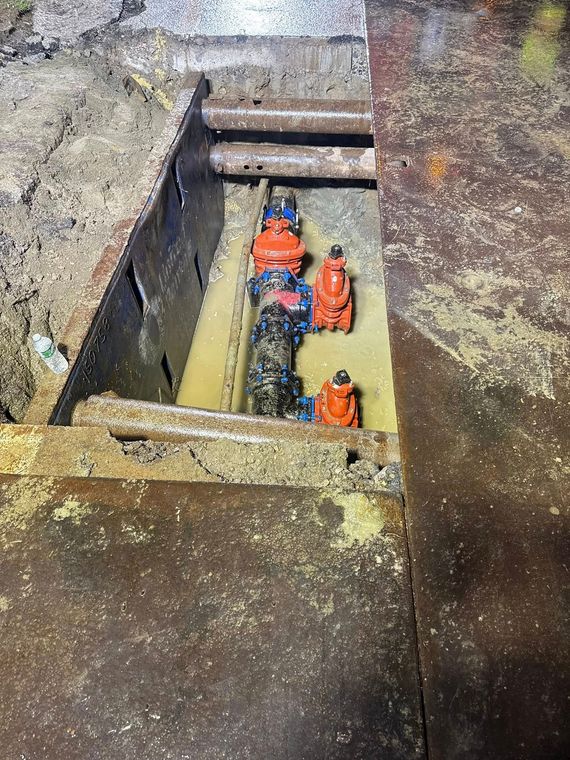 Pipes and valves in a pit, surrounded by metal and dirt. Blue and orange valves are visible.