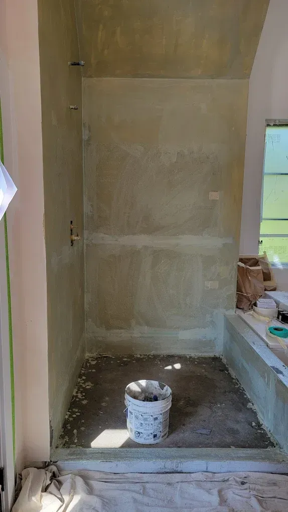 A partially renovated walk-in shower with bare, mudded wall panels, a raw concrete floor, and a bucket sitting in the center.