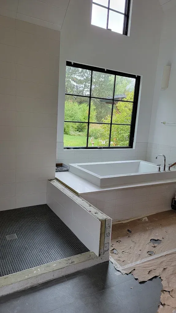 A bathroom under construction with a dark-tiled shower, a white bathtub by a window, and unfinished flooring.