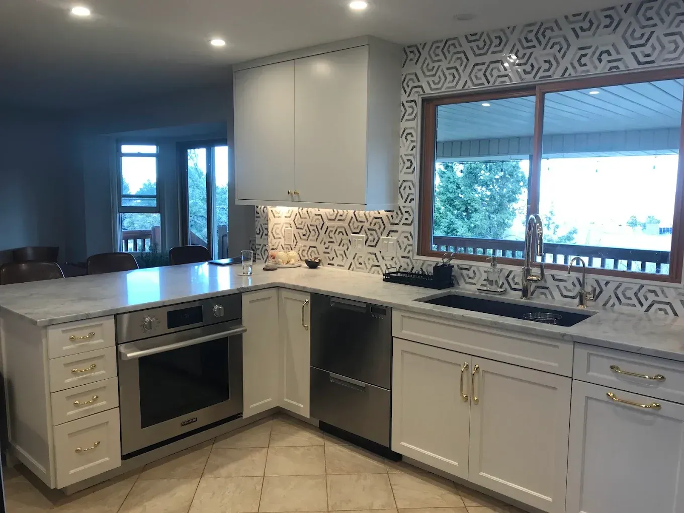 A white kitchen with marble countertops, geometric backsplash tile, stainless steel appliances, and gold hardware.