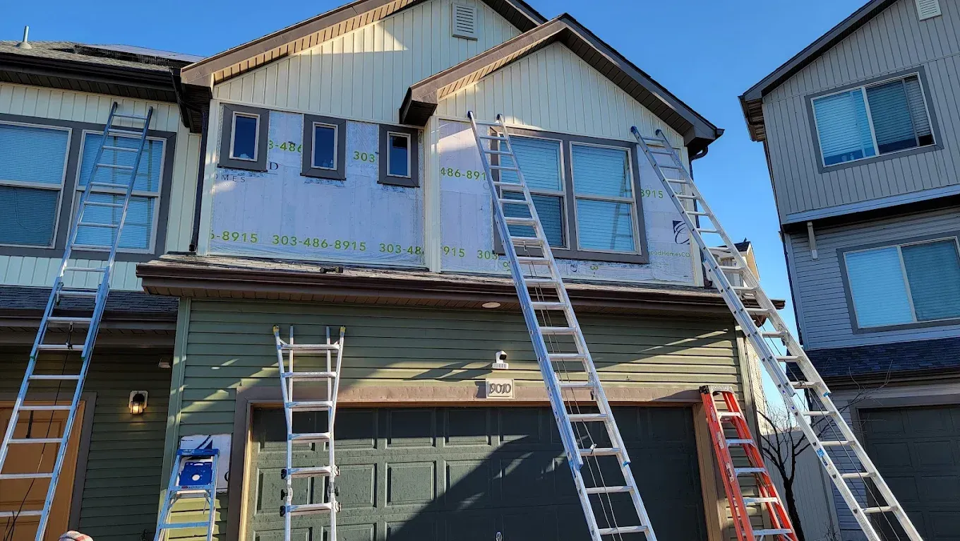Three ladders lean against a house exterior during a siding repair project, with exposed white building wrap visible.