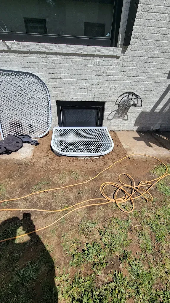 A basement window well with a metal grate cover against a white brick exterior wall.