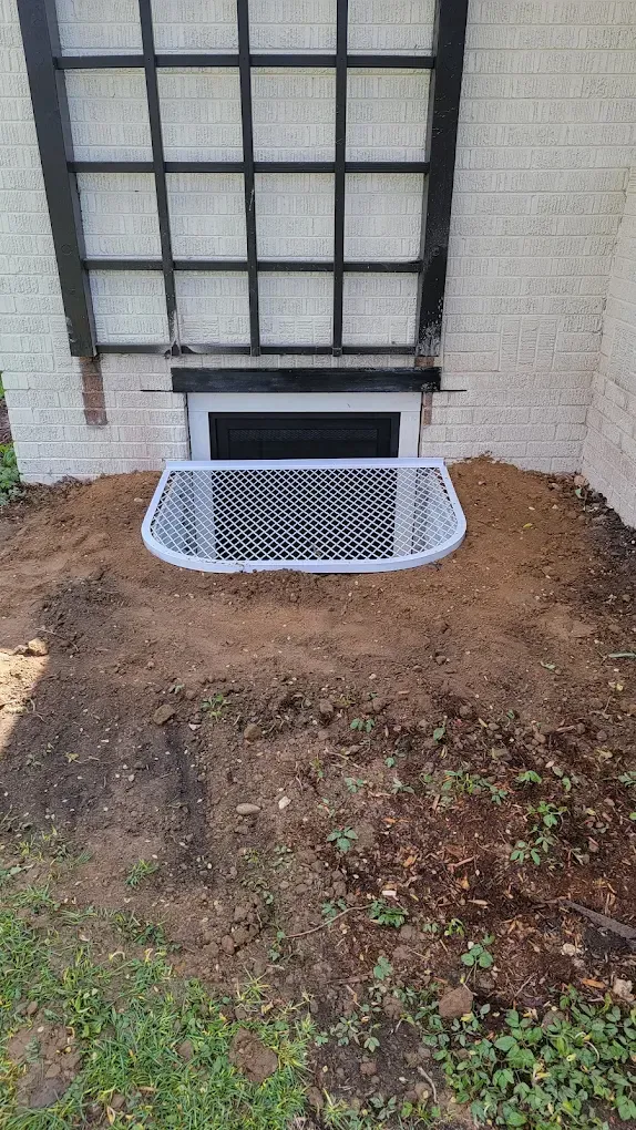 A basement window well with a white metal grate, set into a patch of dirt against a white brick wall with a black trellis.