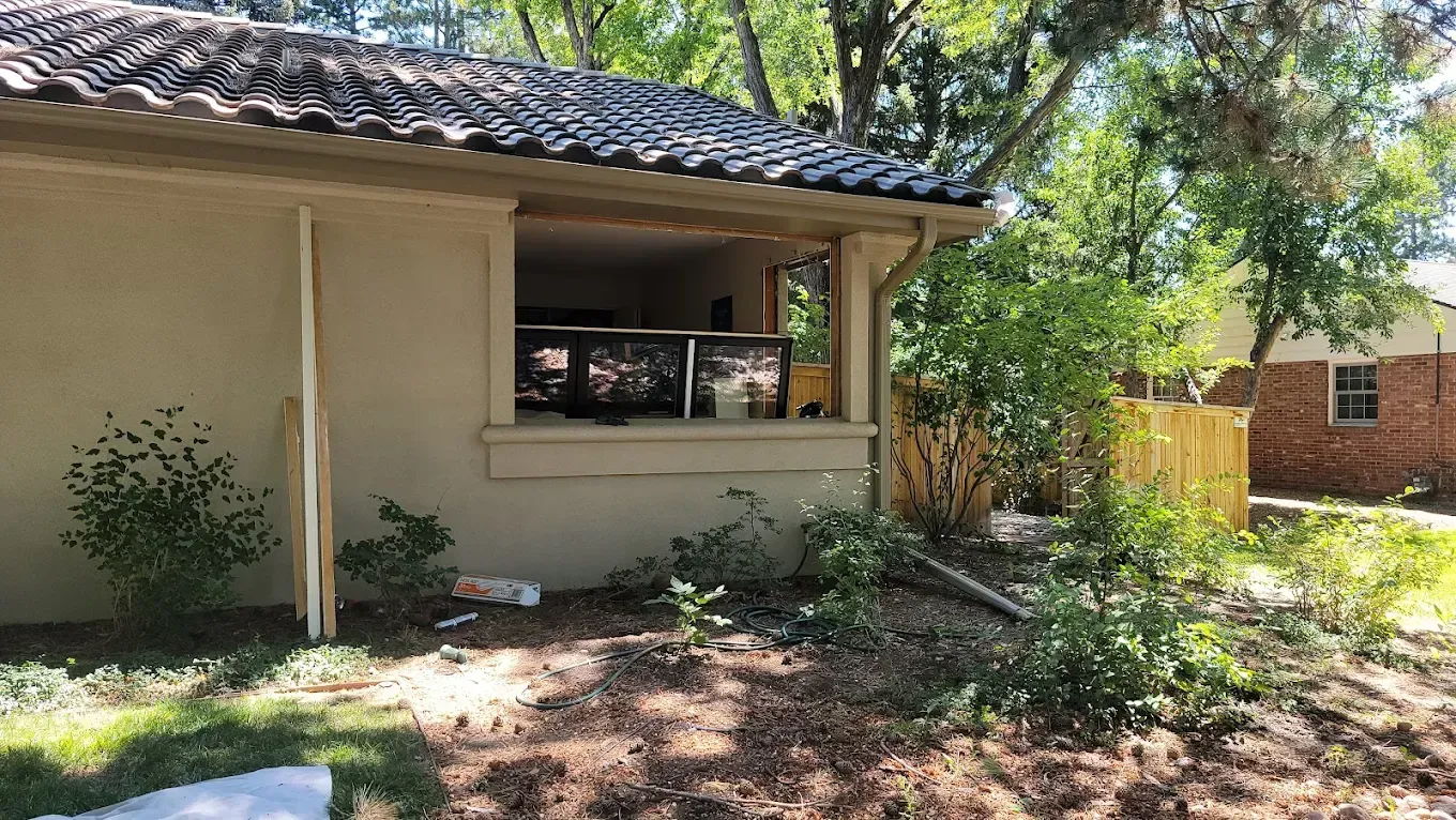 A tan house with a missing window frame, exposing the interior, set in a sunny backyard with trees and a wooden fence.