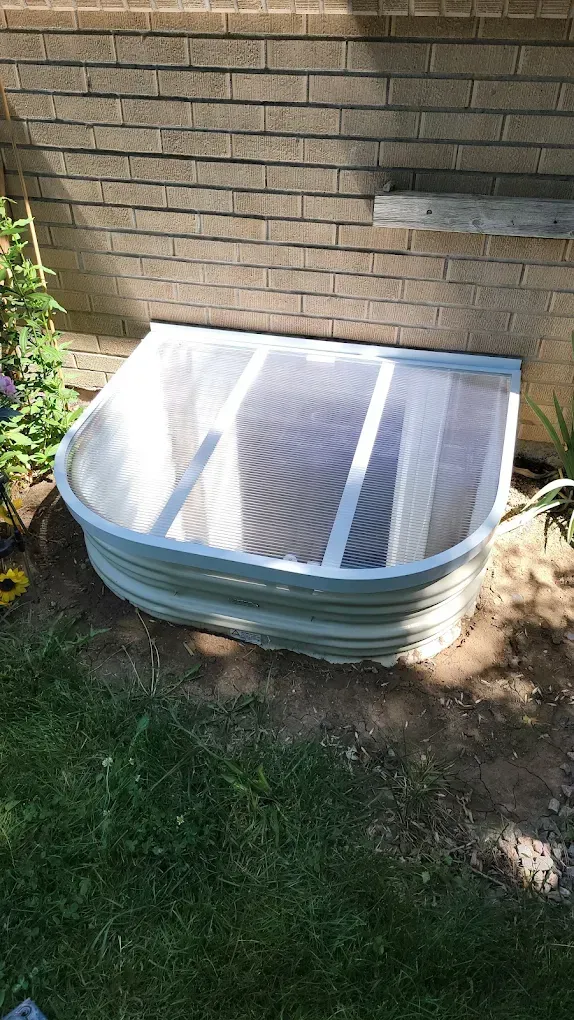 A white window well cover sits over a basement egress window set against a tan brick house wall.