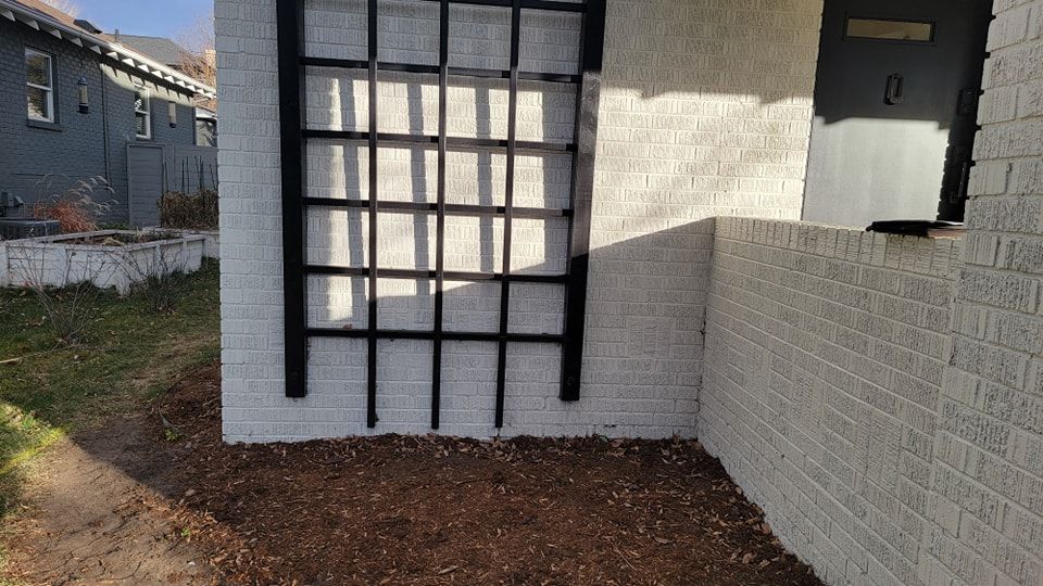 A white brick wall with a black wooden trellis mounted on it, situated in a dirt-covered yard beside a house.