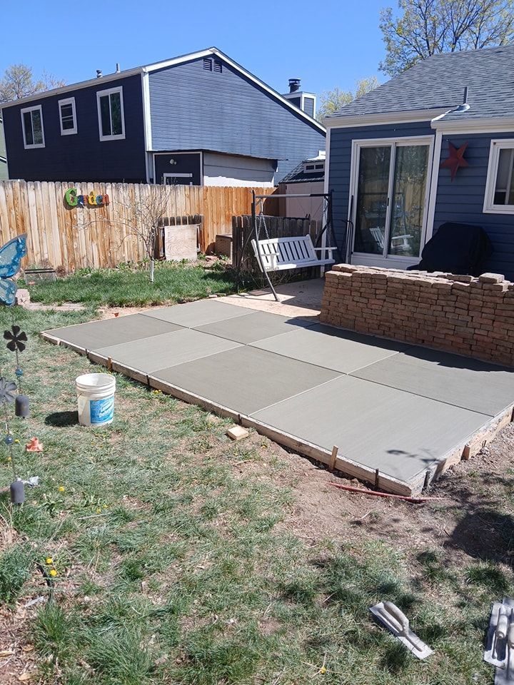Newly poured square concrete patio slabs in a residential backyard next to a blue house and brick retaining wall.